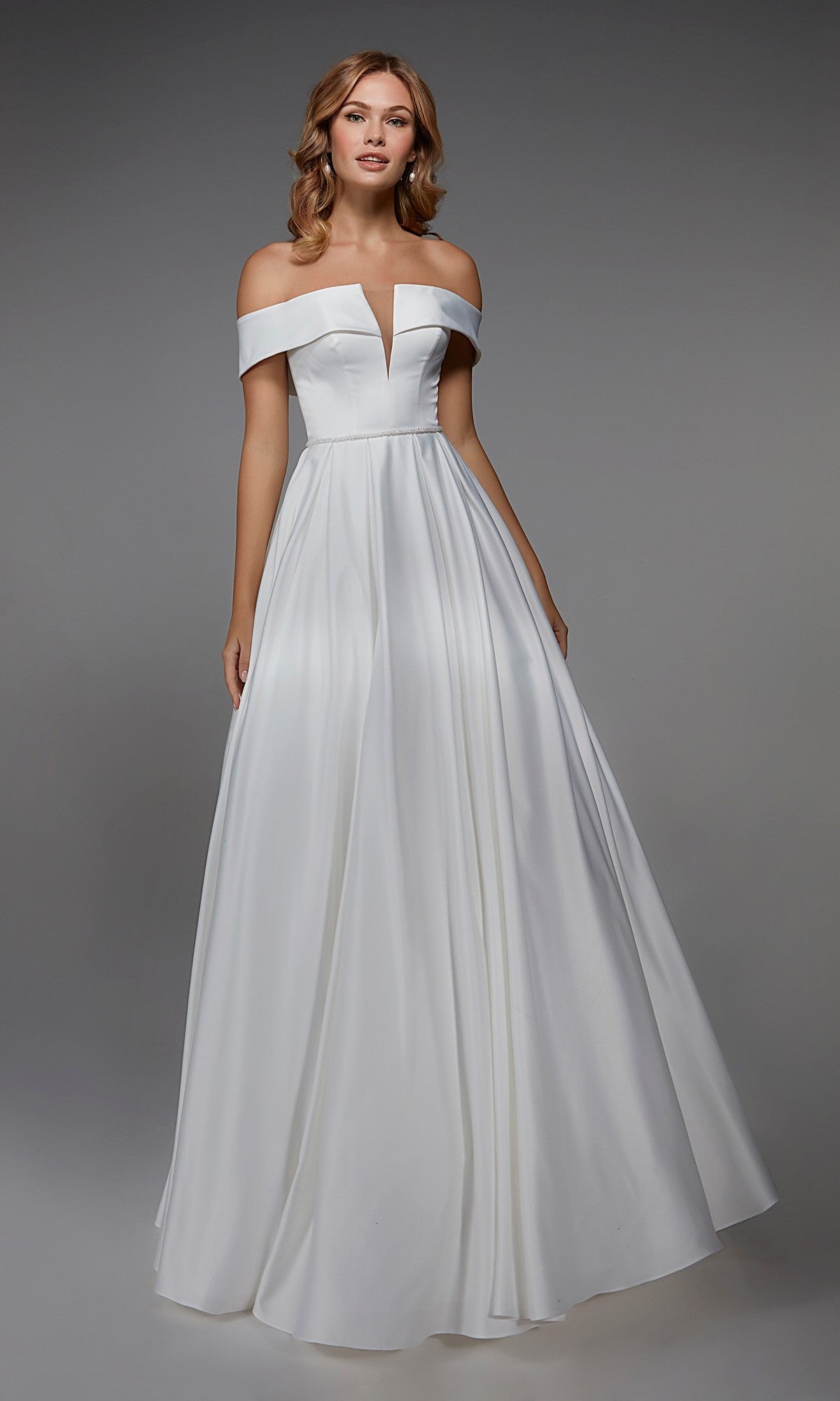 A woman models the Alyce 7023 Long White Dress in Diamond White, featuring an off-the-shoulder fitted bodice and a full, floor-length skirt, standing before a plain gray background.